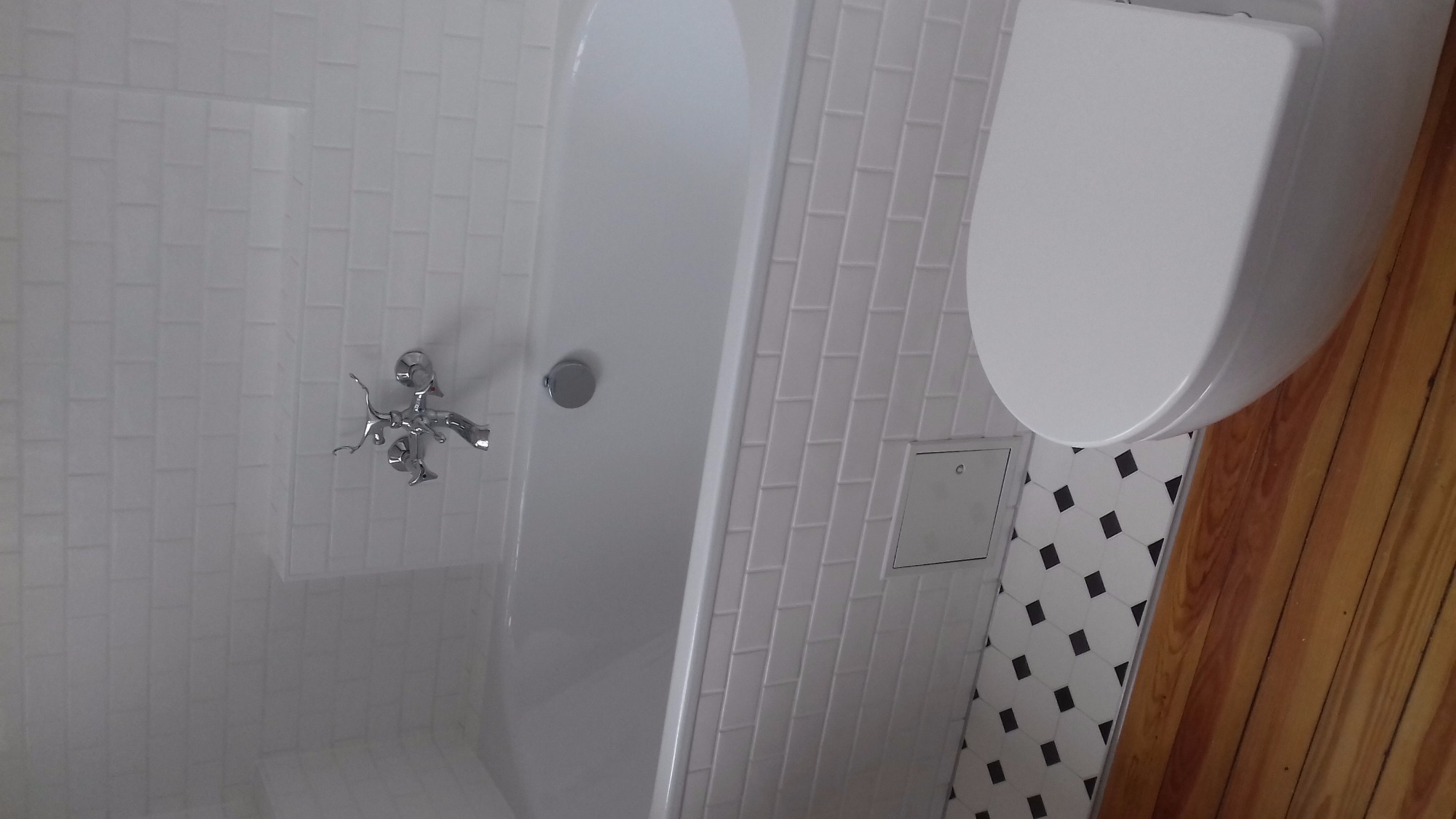 Nowoczesna łazienka z białą wanną, chromowaną armaturą i toaletą, wykończona białymi płytkami typu cegiełka i podłogą w czarno-białe geometryczne wzory, z widocznymi drewnianymi elementami.