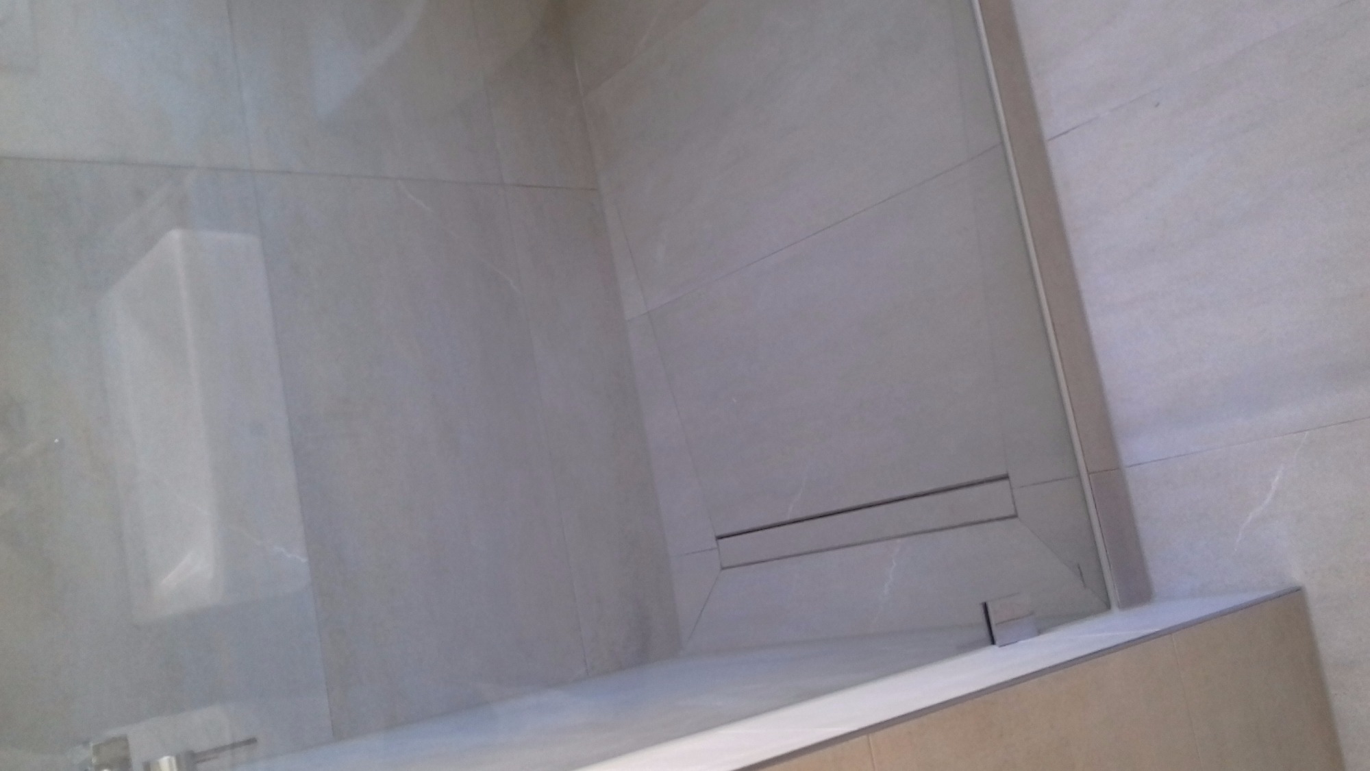 Nowoczesna kabina prysznicowa z odpływem liniowym, wykończona jasnymi, marmurkowymi płytkami ceramicznymi, widoczna szklana ściana z metalowymi okuciami.