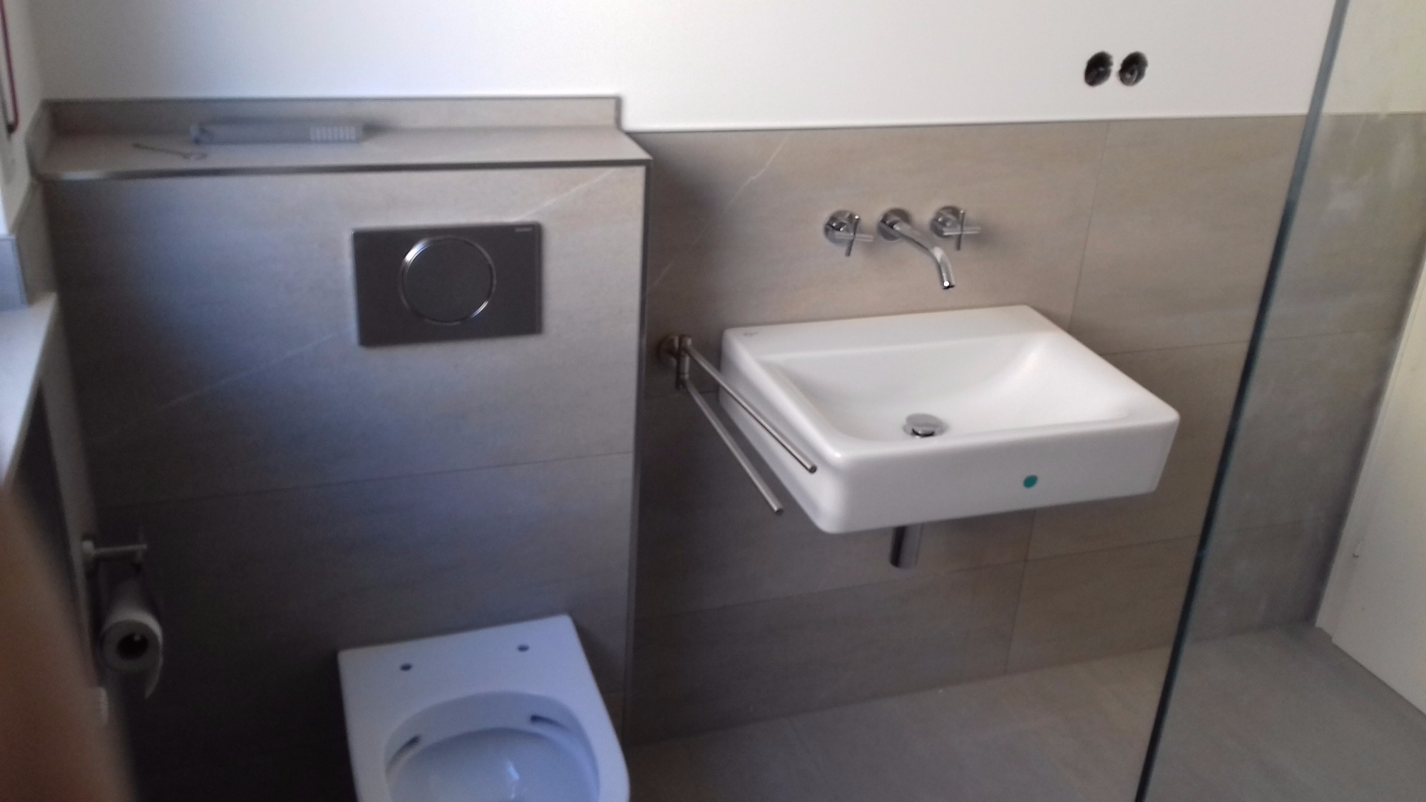 Nowoczesna łazienka z wiszącą białą umywalką, baterią chromowaną, grzejnikiem drabinkowym na ręczniki, toaletą podwieszaną i płytkami w odcieniach beżu.