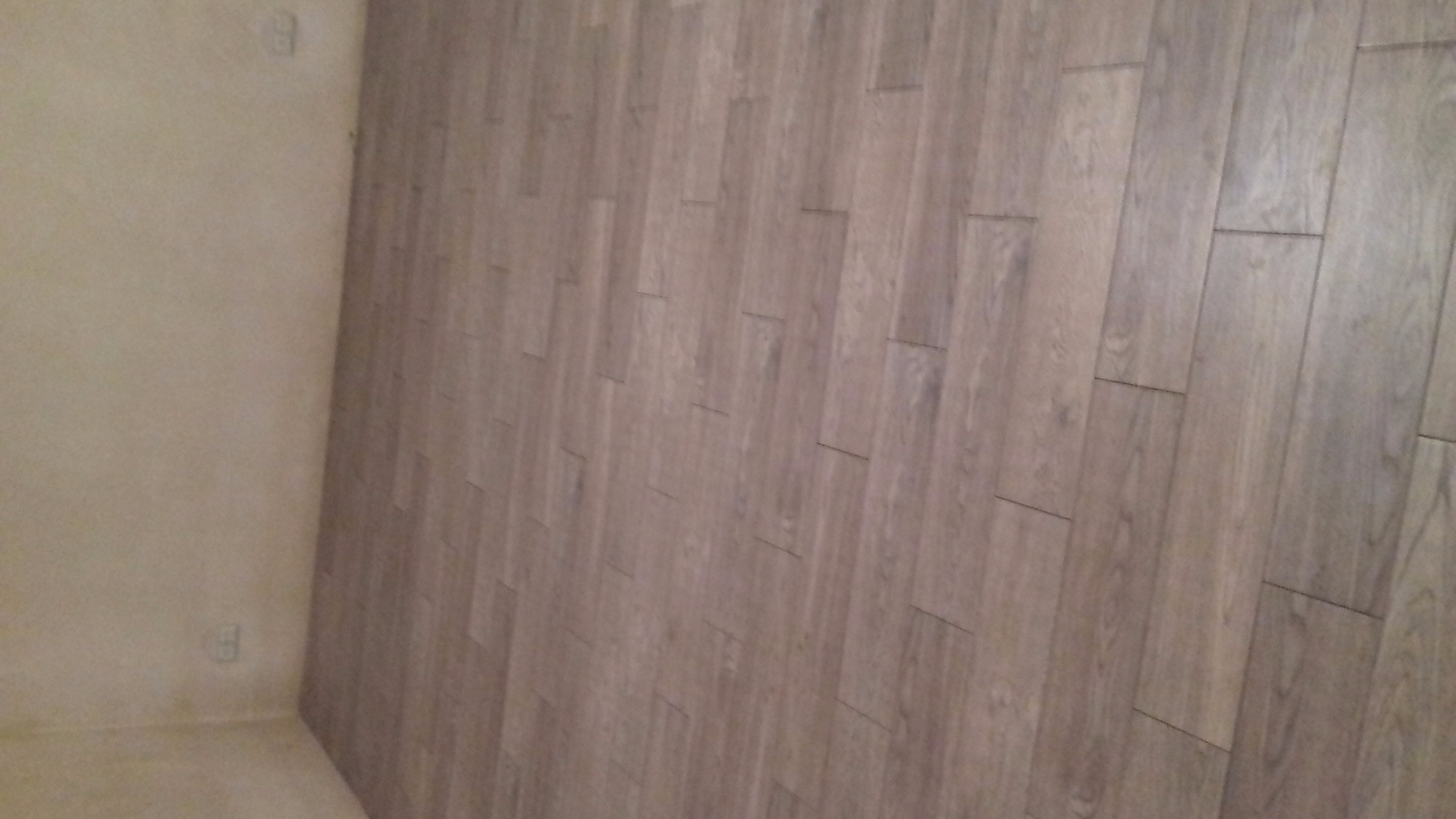 Ukończona ściana z płytek imitujących drewno w odcieniach szarości, widoczna częściowo ściana pomalowana na jasny kolor z gniazdkiem elektrycznym.