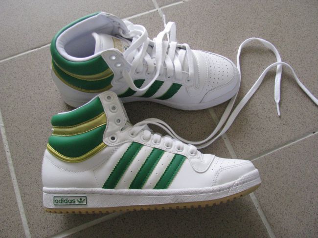 Para białych butów sportowych Adidas Top Ten z zielonymi paskami, stojących na szarej podłodze z płytek. Jeden but jest postawiony za drugim, z rozluźnionymi sznurówkami.