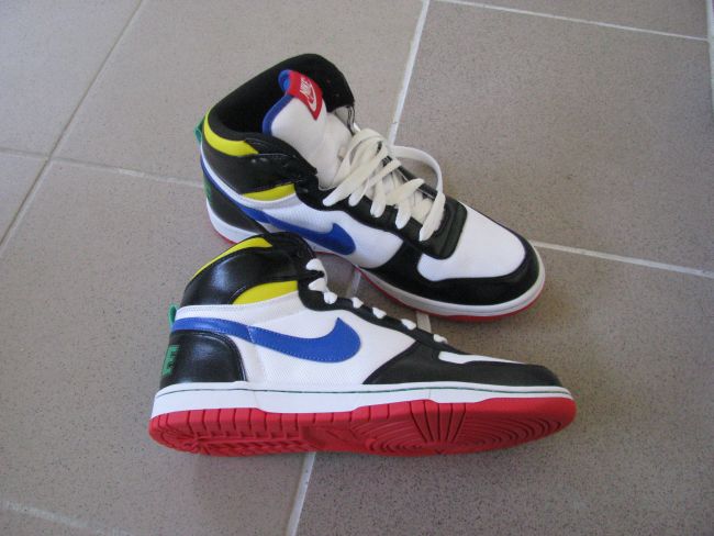 Para sportowych butów typu high-top, biało-czarne z kolorowymi akcentami (żółty, niebieski, czerwony, zielony), leżących na szarej podłodze z płytek. Jeden but leży na drugim, pokazując podeszwę.