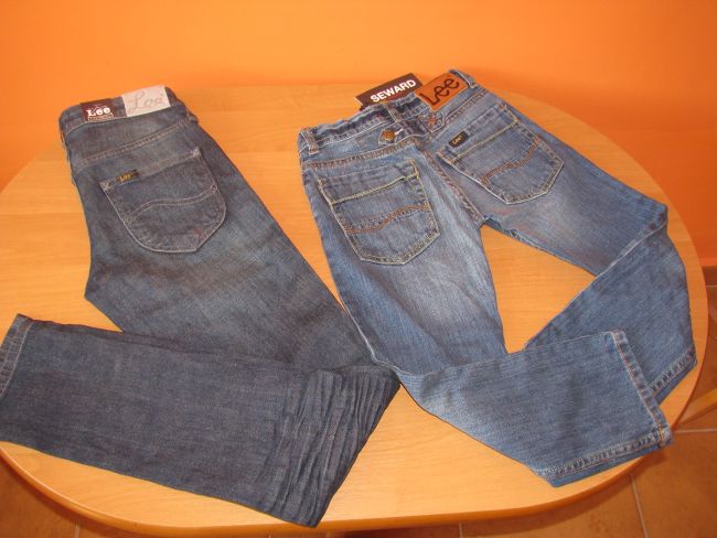 Dwie pary jeansów marki LEE, jedna w ciemniejszym odcieniu, druga jaśniejsza, leżące na okrągłym drewnianym stole na tle pomarańczowej ściany.