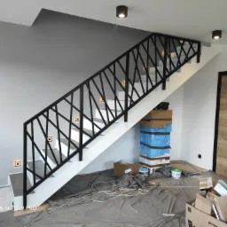 Nowoczesna klatka schodowa z biało-czarną balustradą o geometrycznym wzorze, oświetlona punktowymi lampami ściennymi i sufitowymi, w trakcie prac wykończeniowych z widocznymi materiałami...