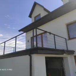 Nowoczesna, minimalistyczna balustrada balkonowa z czarnych, cienkich prętów na tle białej elewacji domu z drewnianą stolarką okienną i błękitnym niebem.