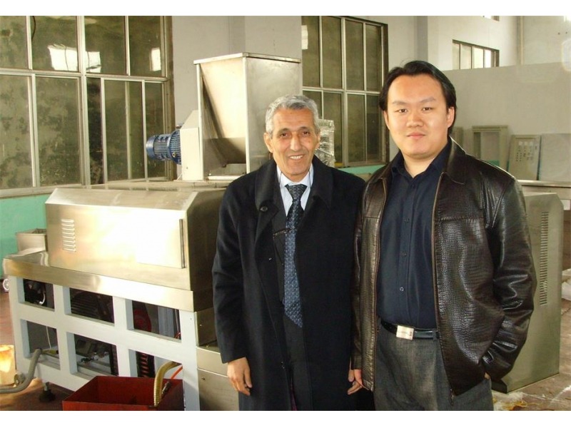 Dwóch mężczyzn pozuje obok przemysłowej maszyny ze stali nierdzewnej z niebieskim silnikiem, widoczne okna i elementy konstrukcyjne w tle.