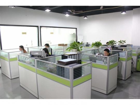 Przestronne biuro z rzędami białych stanowisk pracy oddzielonych szklanymi ściankami z zielonym akcentem, pracownicy przy biurkach, rośliny doniczkowe na niektórych stanowiskach.