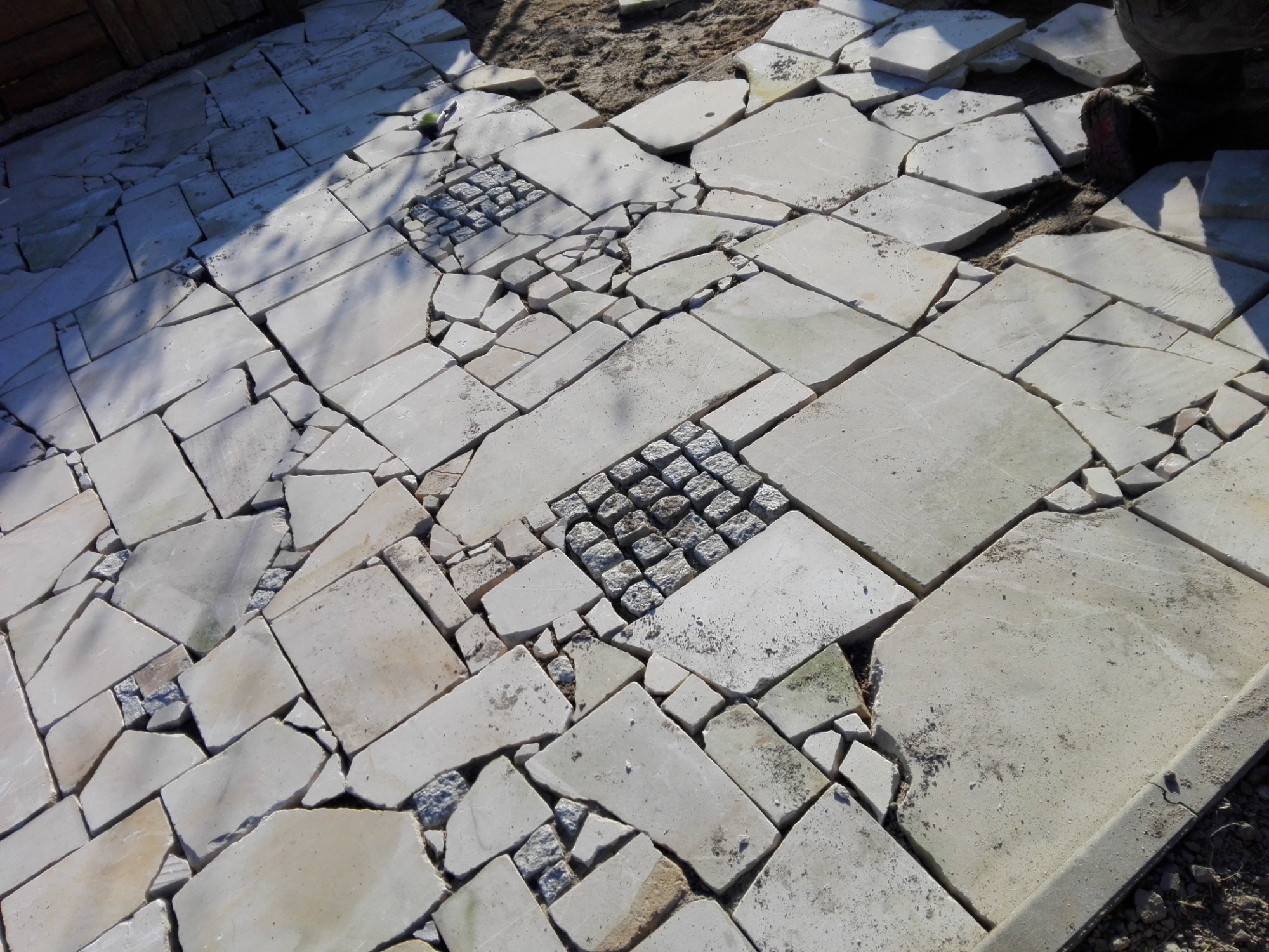 Ułożona nawierzchnia z nieregularnych płyt kamiennych z wkomponowanymi fragmentami kostki brukowej, w trakcie prac wykończeniowych.
