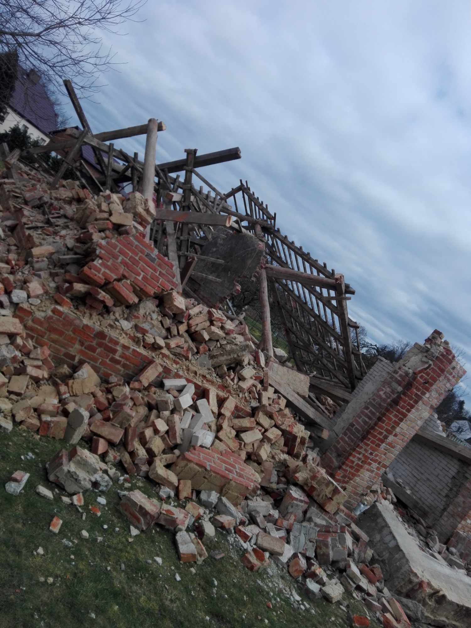 Kupa gruzu i cegieł z drewnianą konstrukcją po zawalonym budynku na trawie, widok na pochmurne niebo.