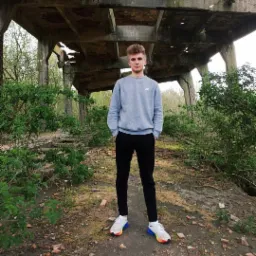 Młody mężczyzna w szarym swetrze i czarnych spodniach stoi na tle zrujnowanej betonowej konstrukcji i zielonej roślinności, prezentując kolorowe buty sportowe.