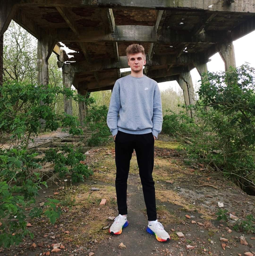 Młody mężczyzna w szarym swetrze i czarnych spodniach stoi na tle zrujnowanej betonowej konstrukcji i zielonej roślinności, prezentując kolorowe buty sportowe.
