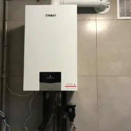 Biały piec gazowy marki Vaillant zamontowany na szarej, kafelkowanej ścianie, z widocznymi rurami doprowadzającymi i odprowadzającymi wodę oraz spalinami.