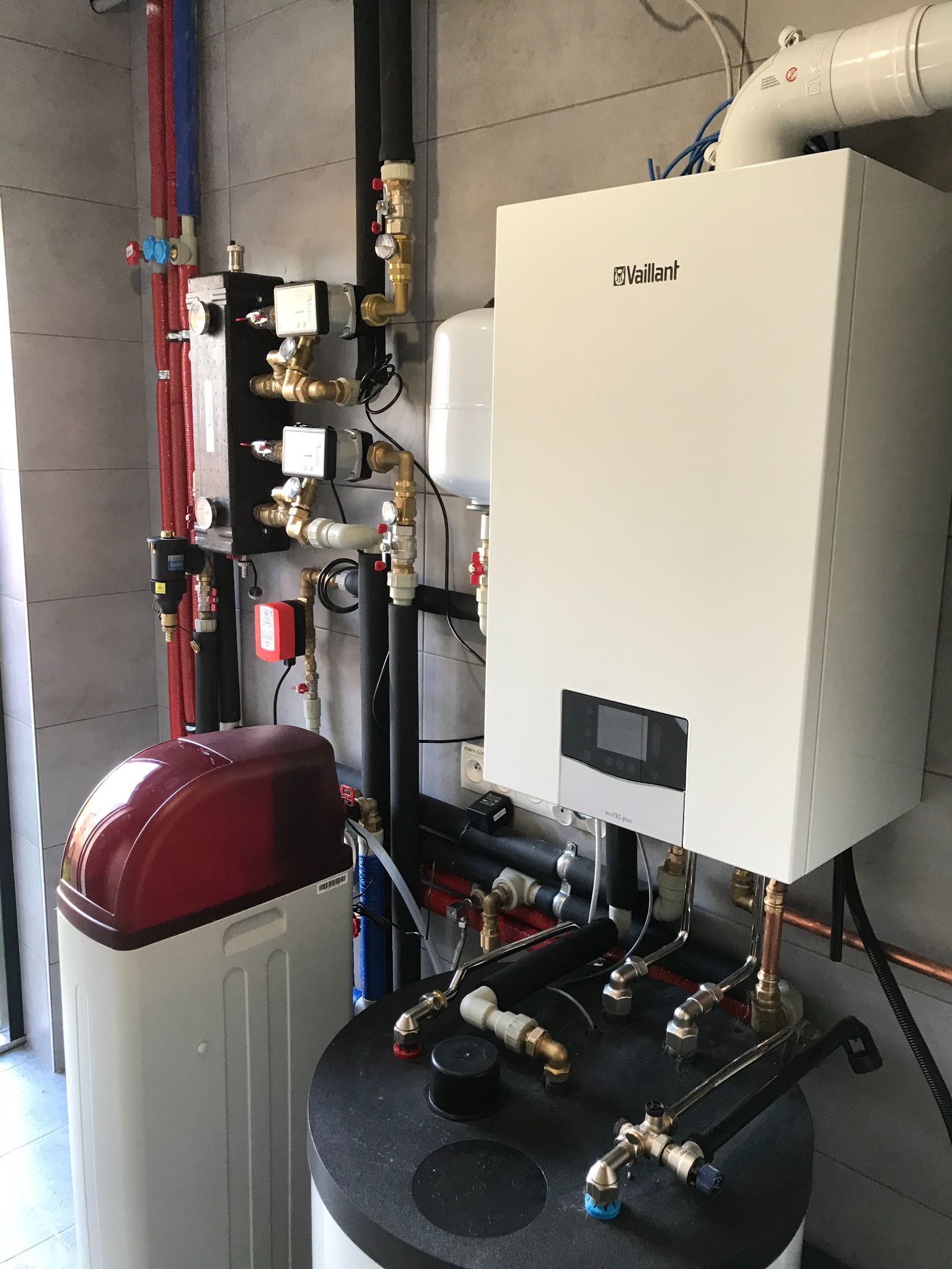 Biały piec gazowy Vaillant z podłączeniami hydraulicznymi, obok zmiękczacz wody z czerwoną pokrywą i czarnym filtrem, widoczne rury miedziane i zawory, szare płytki na ścianie.
