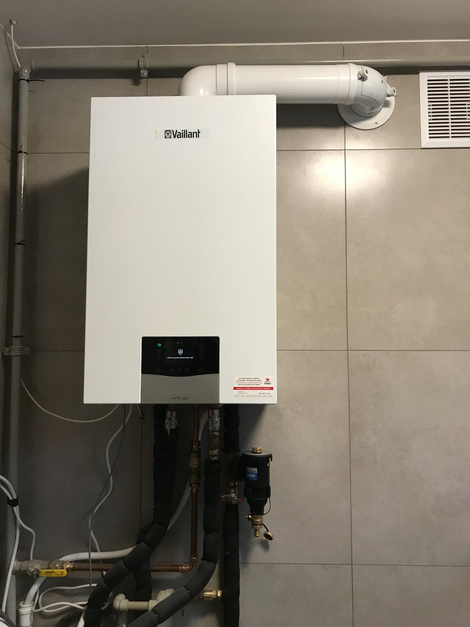 Biały piec gazowy marki Vaillant zamontowany na szarej, kafelkowanej ścianie, z widocznymi rurami doprowadzającymi i odprowadzającymi wodę oraz spalinami.