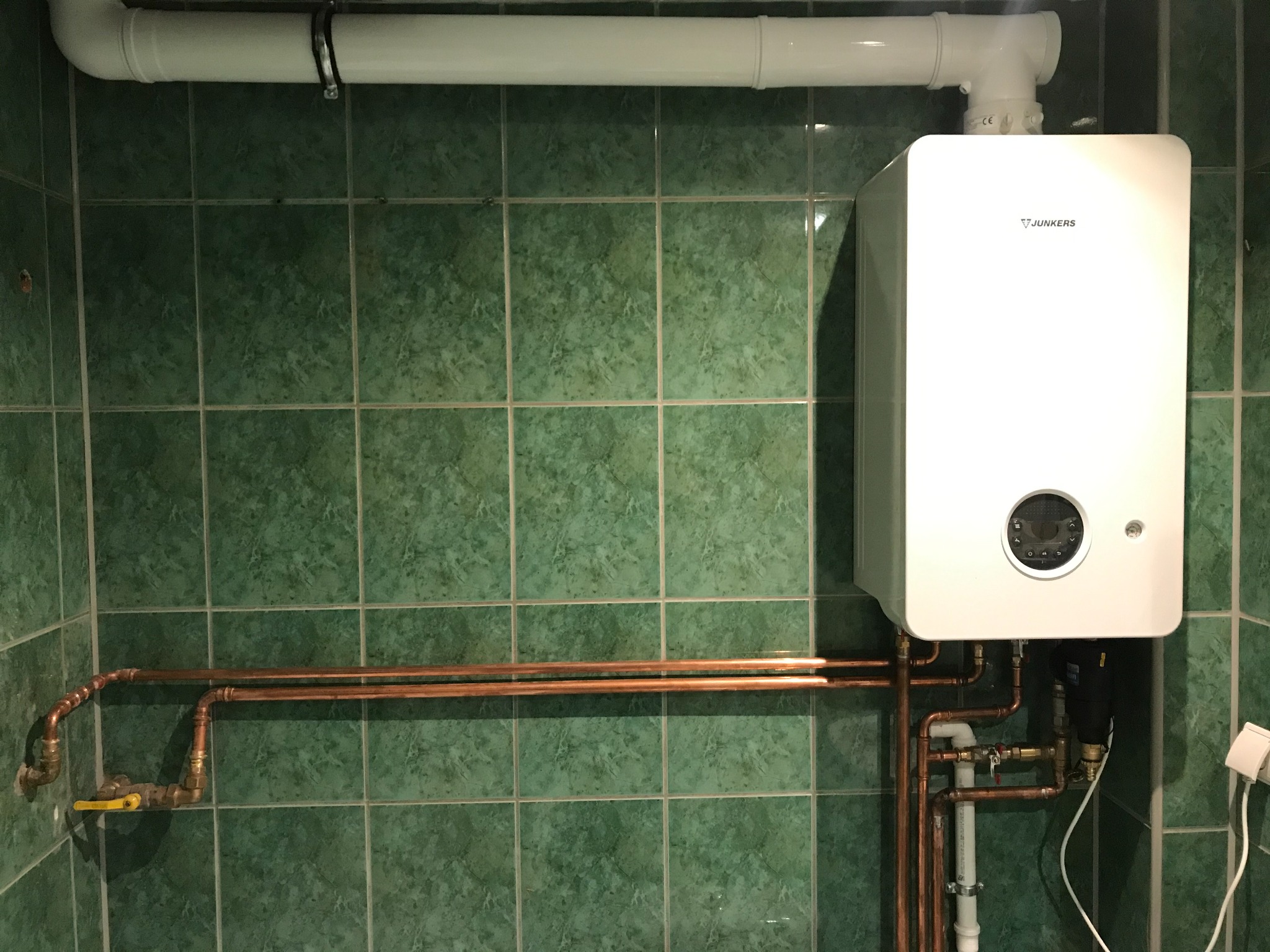 Biały piec gazowy Junkers zamontowany na zielonej, kwadratowej ścianie z widocznymi miedzianymi rurami i białym przewodem wentylacyjnym.