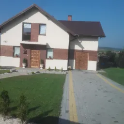 Dwukondygnacyjny dom jednorodzinny z brązowym dachem, kremową elewacją z brązowymi akcentami, drewnianymi drzwiami wejściowymi i numerem domu 425, otoczony trawnikiem, ozdobnymi krzewami...