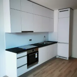 Nowoczesna, minimalistyczna kuchnia z białymi szafkami i czarnym blatem, z widocznymi elementami wykończeniowymi i drewnianą podłogą.