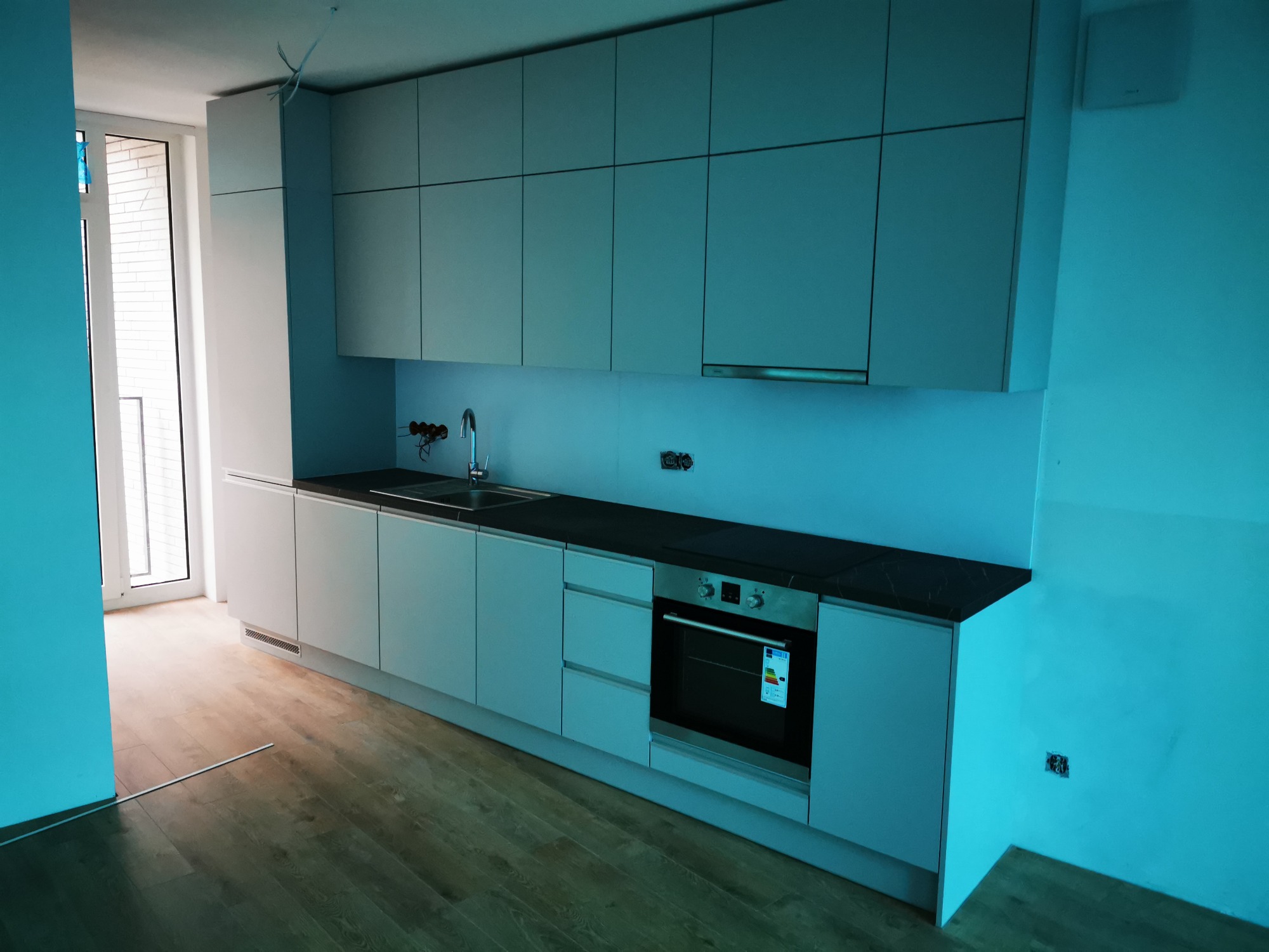 Nowoczesna, minimalistyczna zabudowa kuchenna w odcieniach szarości z czarnym blatem, widok na fragment okna i drewnianą podłogę, w trakcie montażu instalacji elektrycznej.