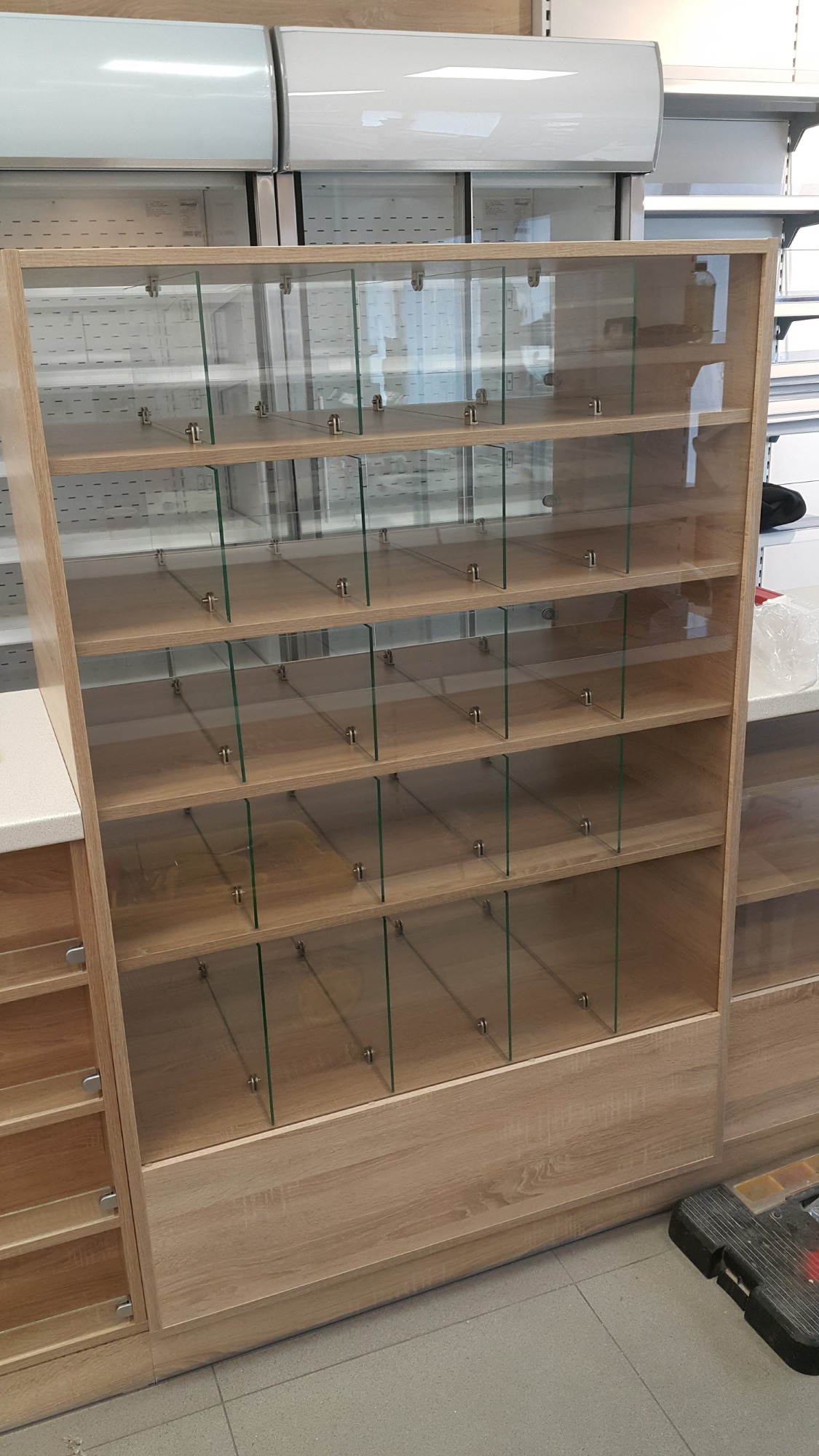 Pusta, jasna, drewniana witryna sklepowa z szklanymi półkami i przegródkami, widoczna skrzynka z narzędziami w prawym dolnym rogu.