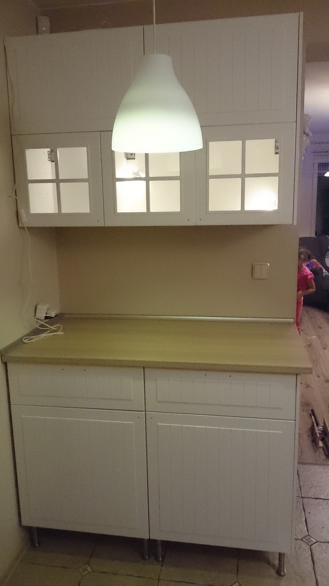 Wykonana na wymiar biała szafka kuchenna z przeszklonymi górnymi szafkami i blatem w odcieniach beżu, oświetlona lampą wiszącą, zrealizowana w Łodzi.