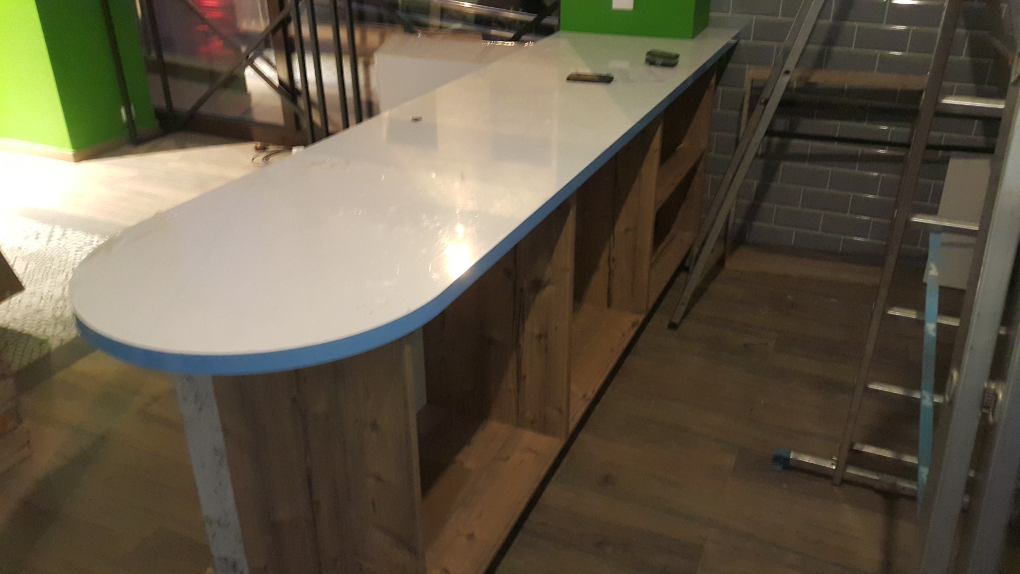 Biały blat baru z zaokrąglonymi krawędziami i niebieskim obramowaniem, wsparty na drewnianej konstrukcji z otwartymi półkami, w tle drabina oparta o ścianę z szarych płytek.