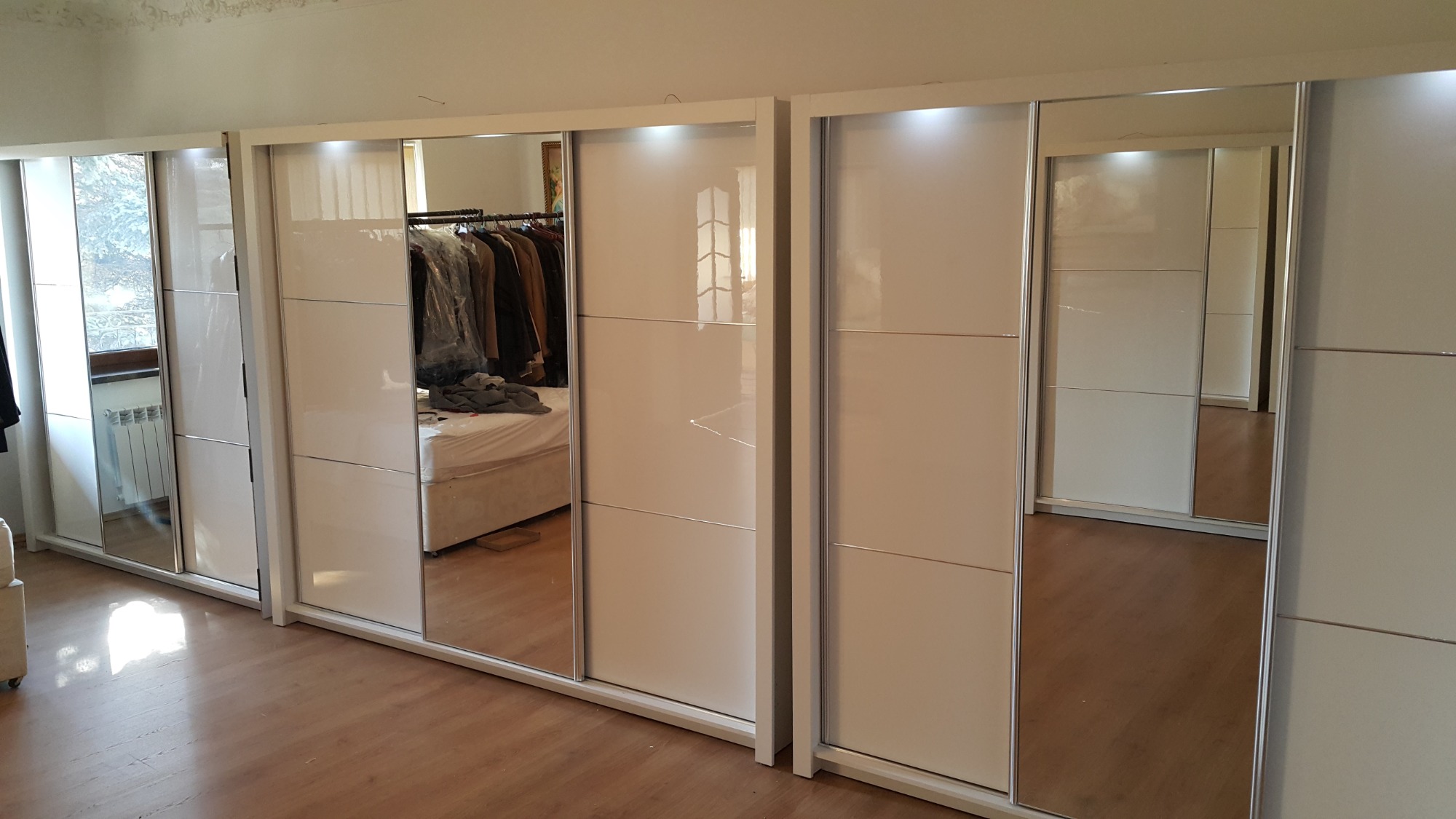 Biała szafa przesuwna z lustrami w sypialni, widoczne ubrania na wieszakach odbijające się w lustrze.