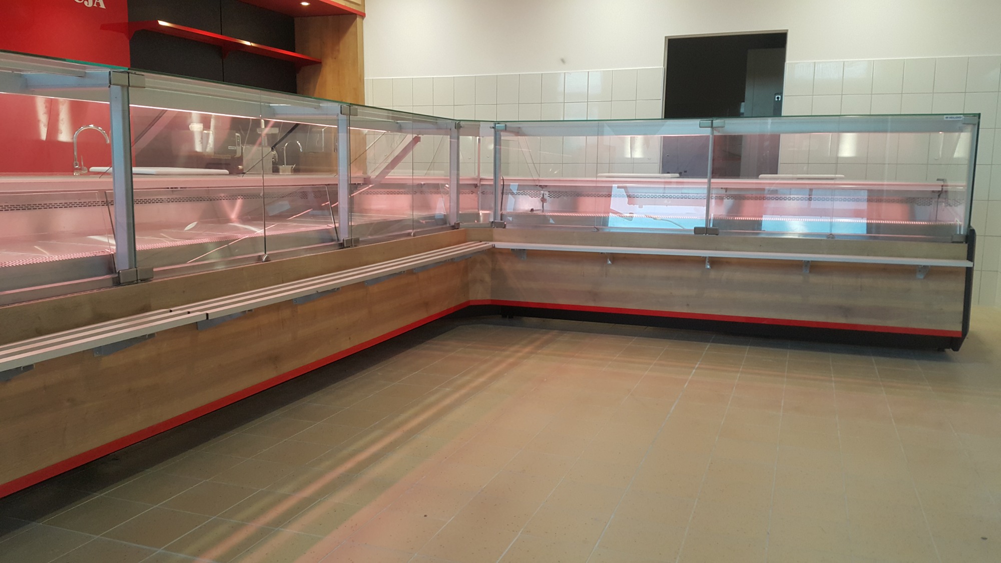 Pusta, narożna lada chłodnicza z drewnianą obudową i szklaną witryną, z widocznymi elementami oświetlenia wewnętrznego i umywalką w tle, na tle wyłożonej kafelkami ściany.