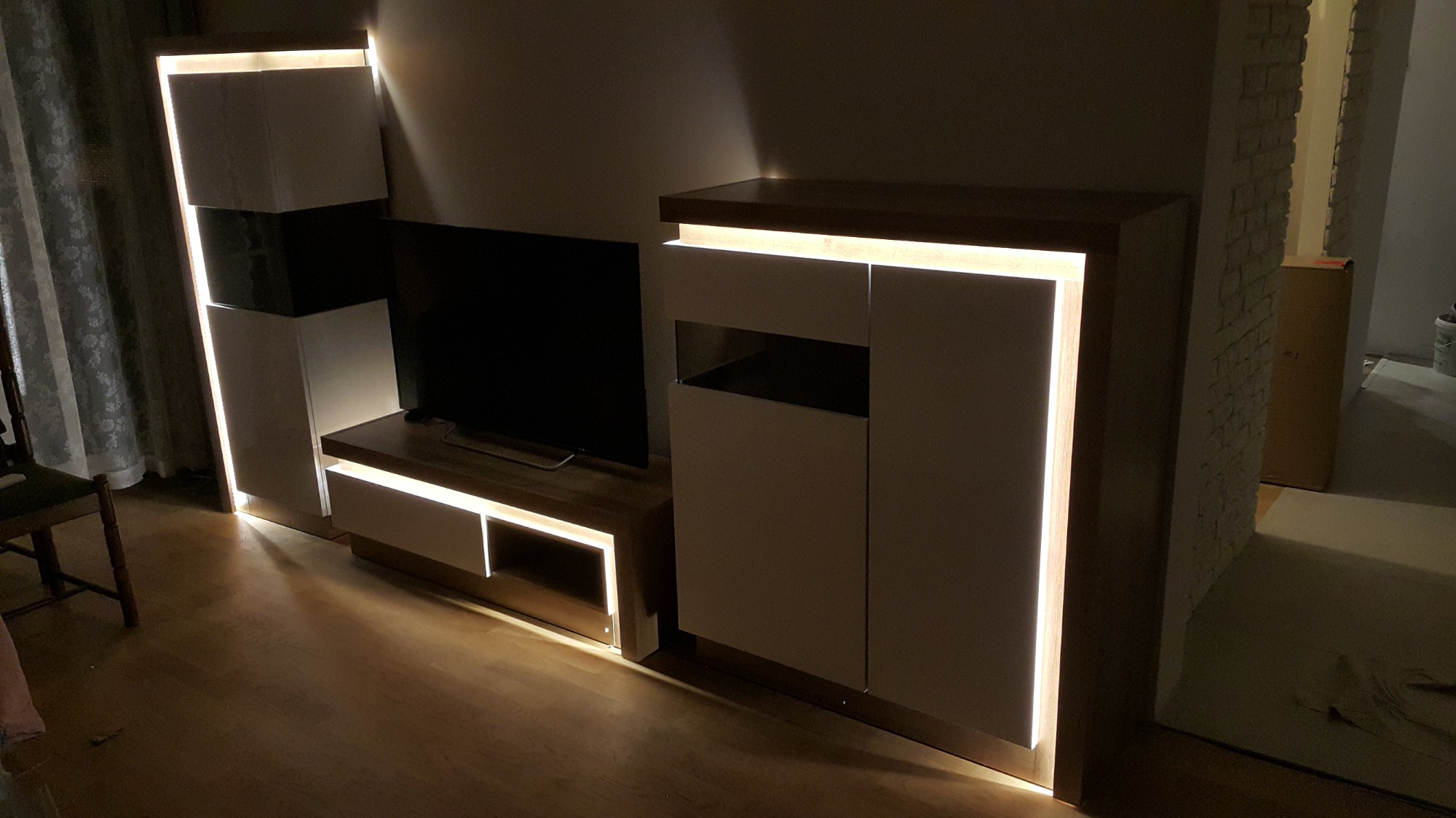 Nowoczesna meblościanka z podświetleniem LED, obejmująca szafki i półkę pod telewizor, ustawiona w salonie z drewnianą podłogą i widocznymi zasłonami po lewej stronie.