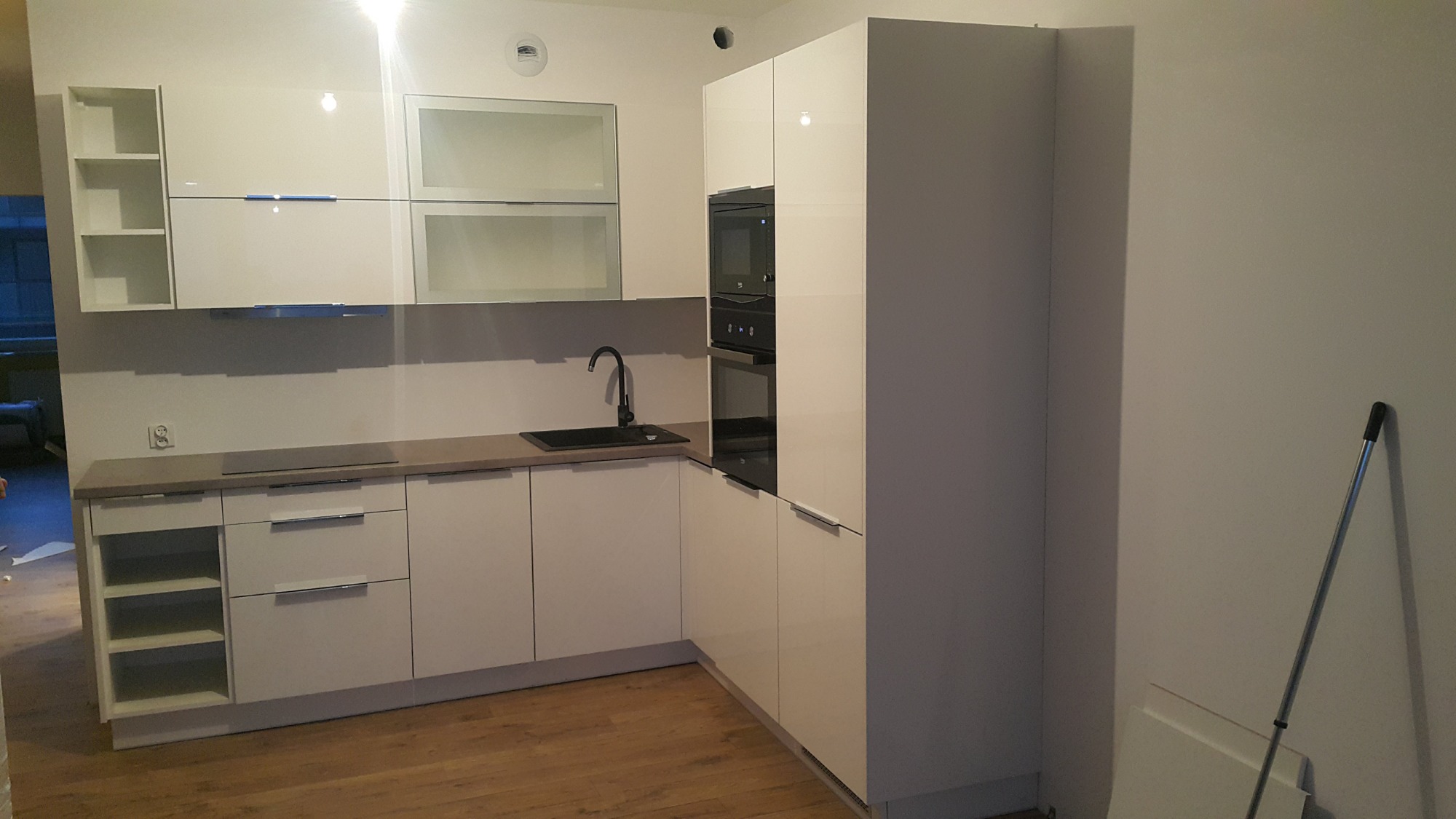 Nowoczesna, jasna kuchnia z białymi, lakierowanymi frontami szafek, czarną armaturą i drewnianą podłogą. W rogu oparta o ścianę rura od odkurzacza.