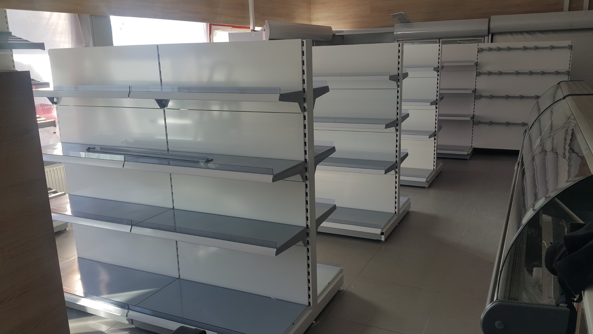 Białe, metalowe regały sklepowe z szarymi półkami, ustawione w rzędach w pustym pomieszczeniu, widoczne z perspektywy pod lekkim kątem.