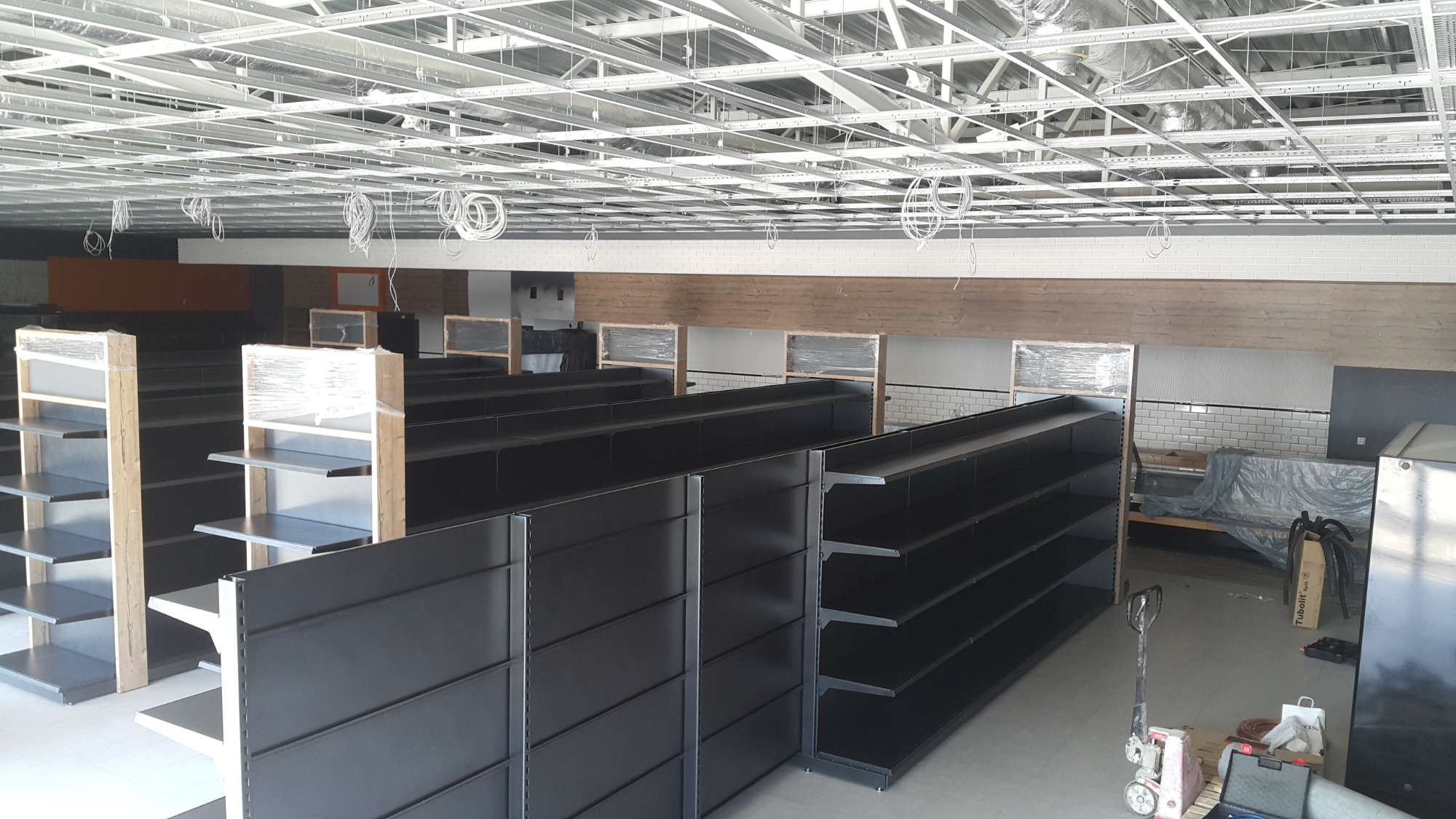 Montaż czarnych regałów sklepowych w surowym wnętrzu z widoczną instalacją pod sufitem, częściowo zabezpieczone folią.