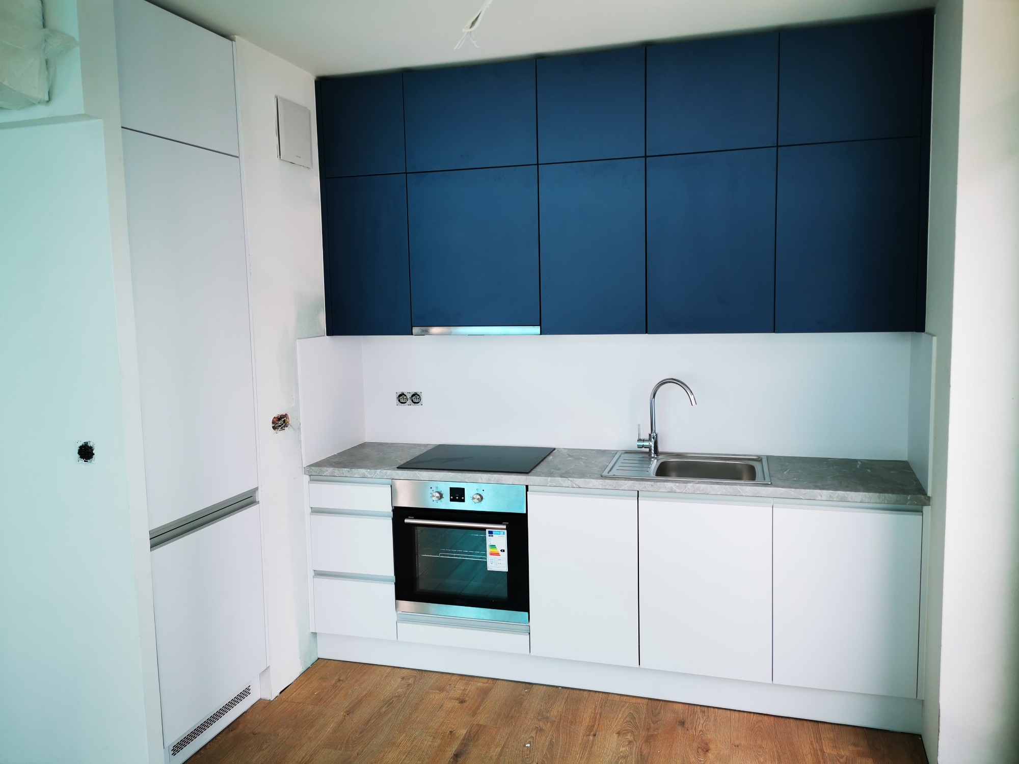 Nowoczesna kuchnia z białymi frontami szafek i granatowymi górnymi szafkami, szary blat, zlewozmywak ze stali nierdzewnej i piekarnik z etykietą energetyczną, widoczne niedokończone prace...