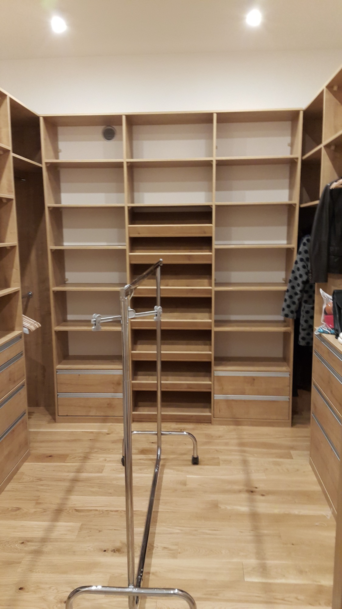 Pusta, drewniana garderoba na wymiar z szufladami i półkami, widok z przodu, na podłodze jasne panele, w tle widoczna kratka wentylacyjna.