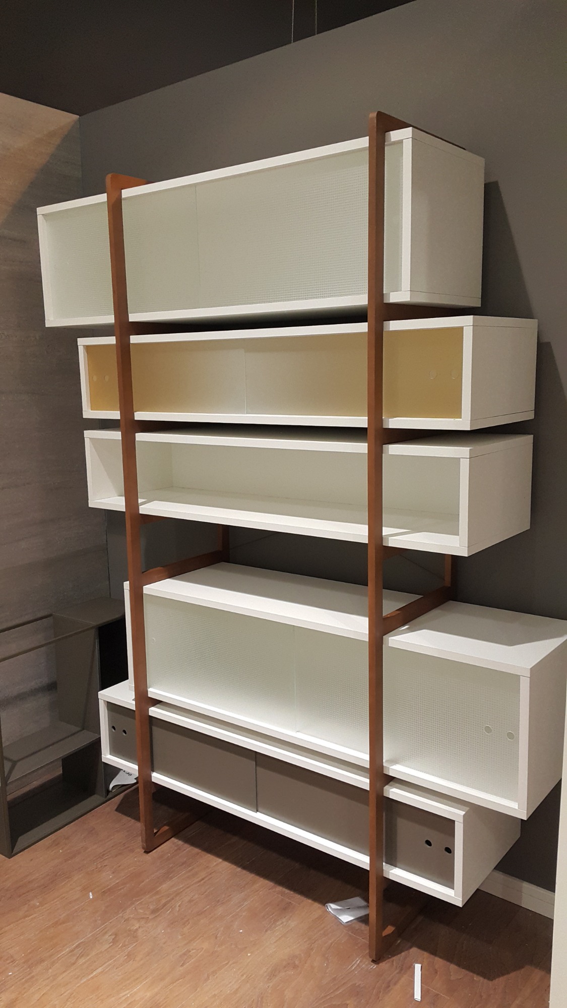 Regał modułowy z białymi szafkami i półkami, wsparty na drewnianej konstrukcji, na tle szarej ściany i drewnianej podłogi.