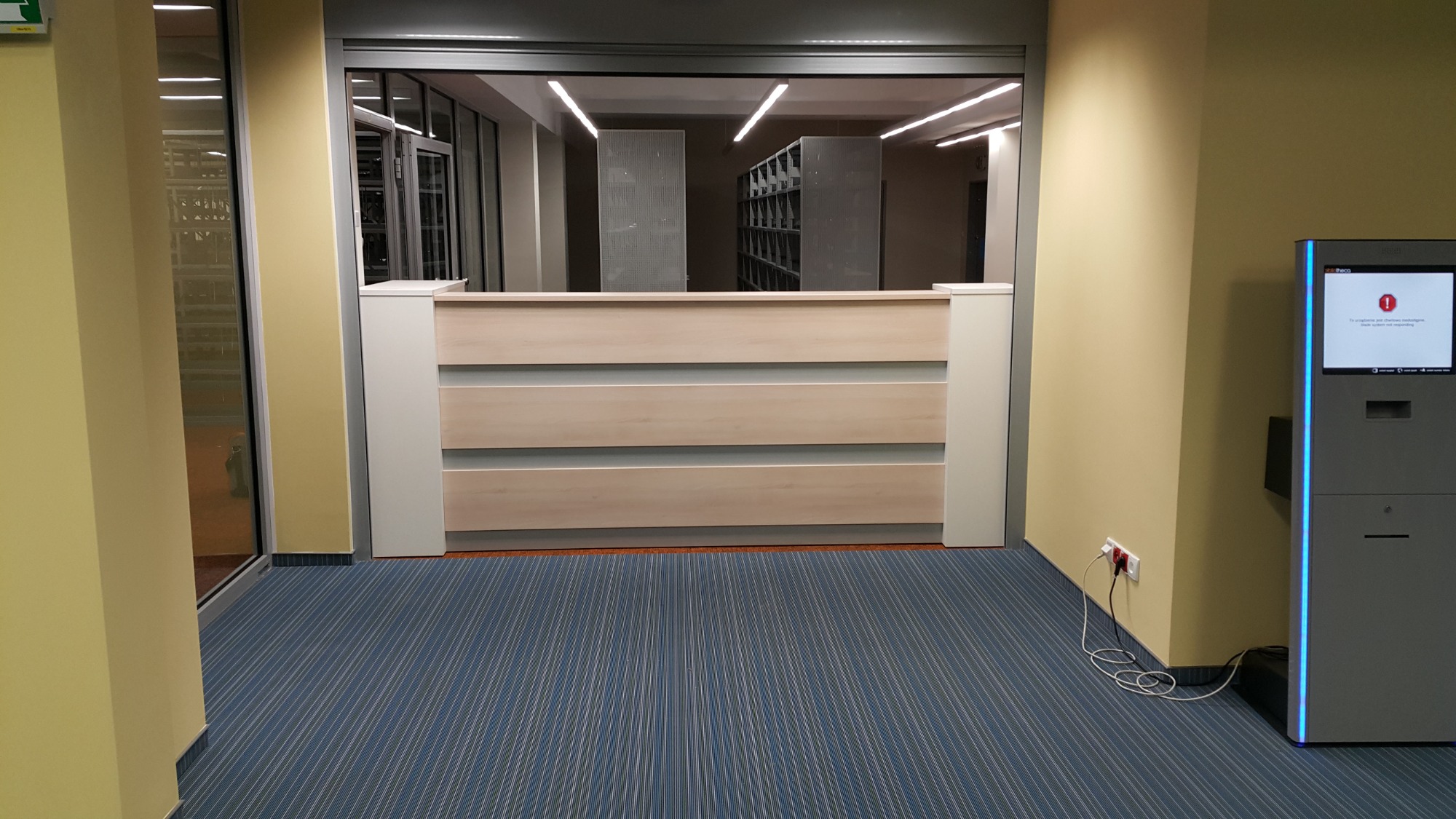 Jasna, drewniana lada recepcyjna z trzema poziomymi panelami, widziana z perspektywy niskiego kąta, z automatem samoobsługowym po prawej stronie i regałami w tle.