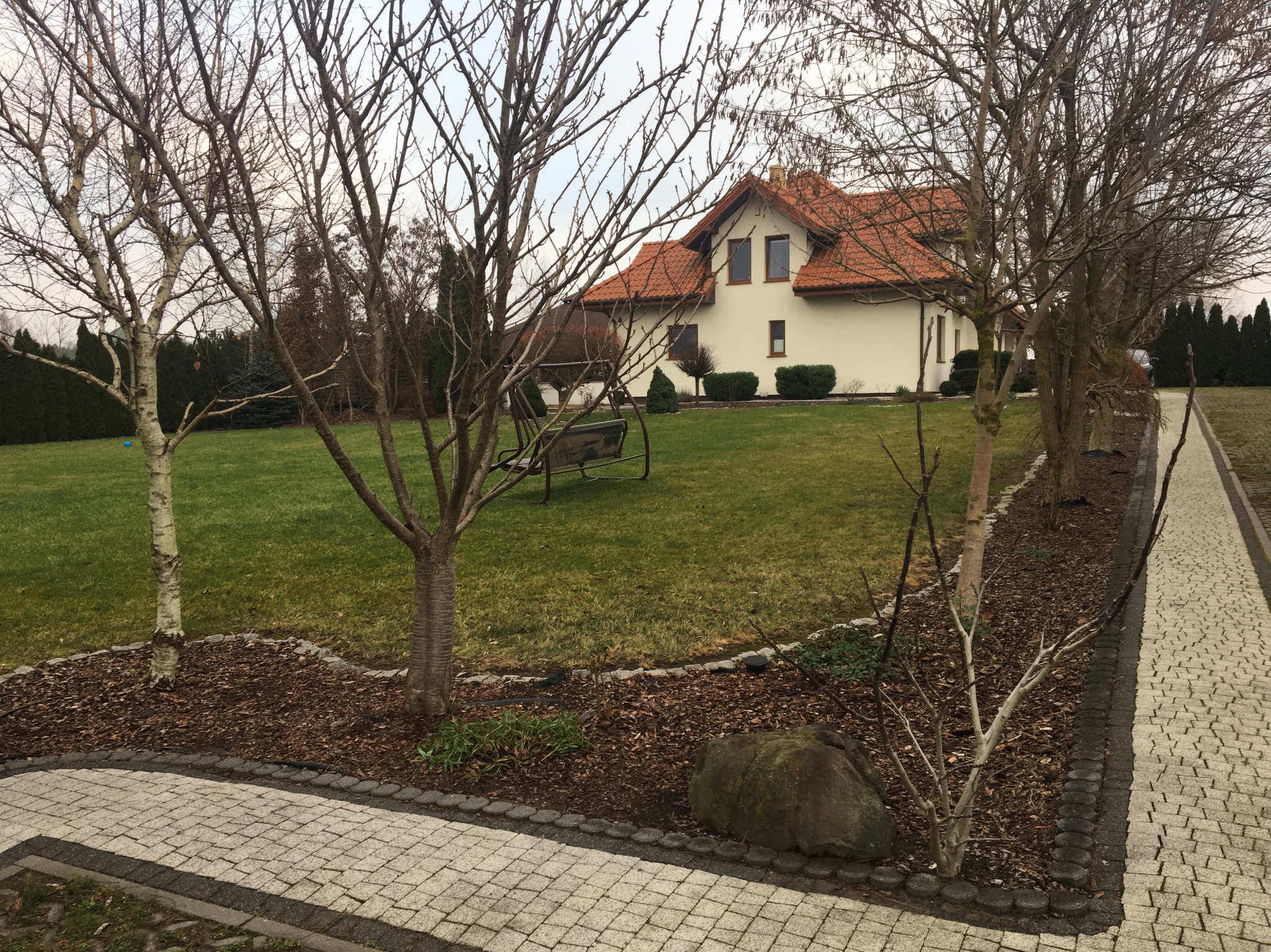 Dom jednorodzinny z czerwonym dachem widziany z perspektywy ogrodu z kostki brukowej i drzew bez liści w pochmurny dzień.