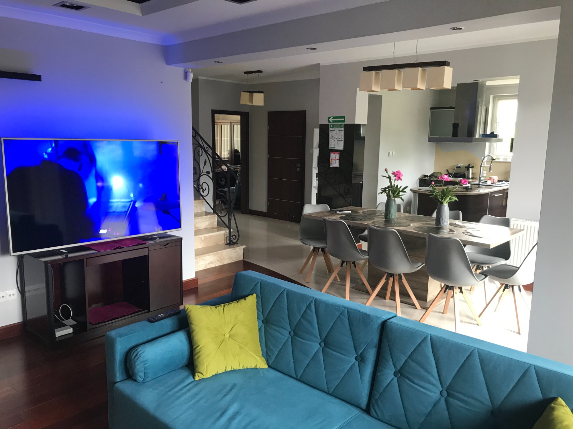 Jasne wnętrze domu z turkusową sofą, telewizorem z niebieskim ekranem, stołem jadalnym z szarymi krzesłami i bukietami różowych kwiatów w wazonach.