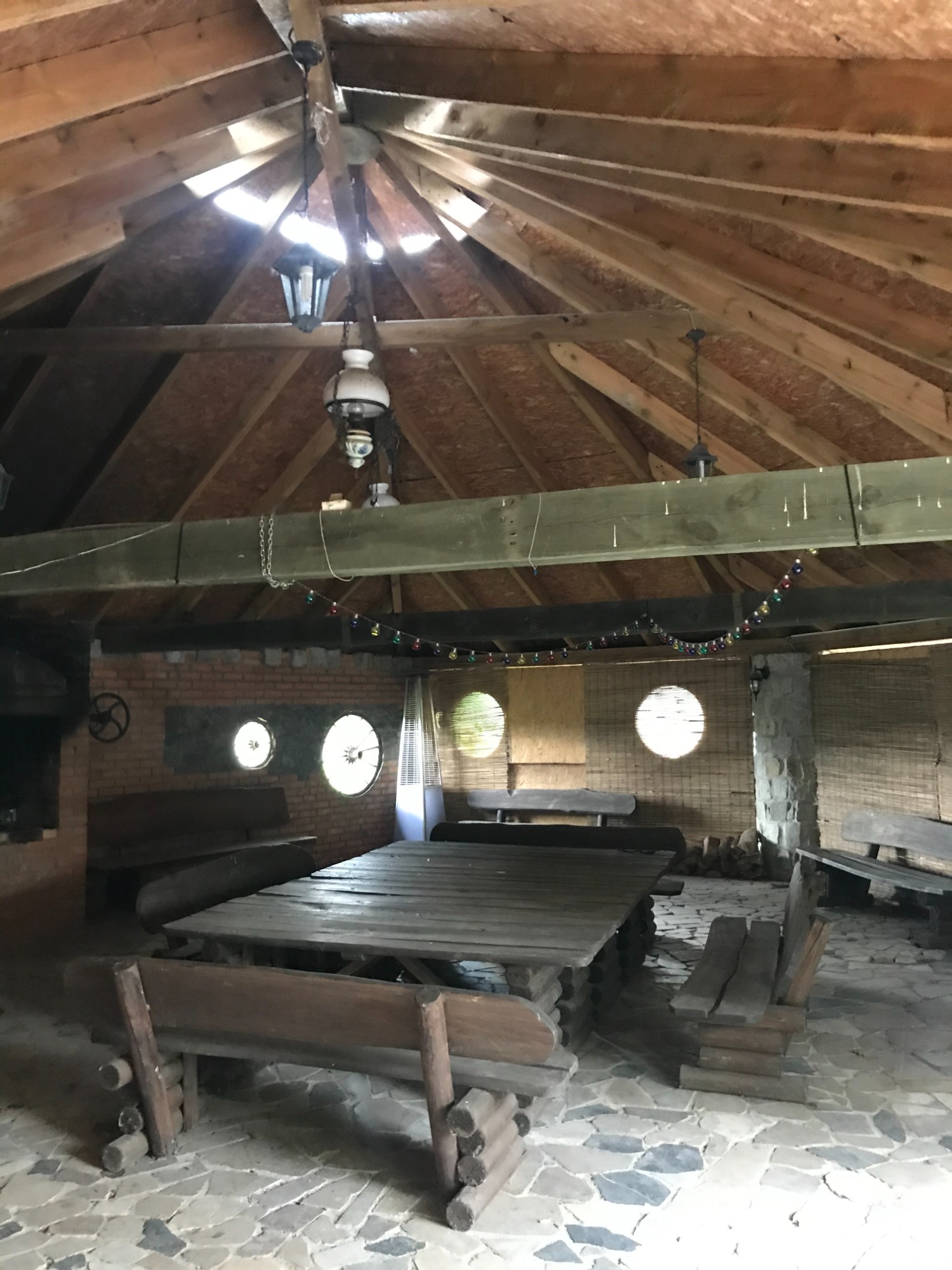 Rustykalna altana z drewnianym dachem, kamienną podłogą i masywnym stołem z ławkami, oświetlona wiszącymi lampami i girlandą żarówek.