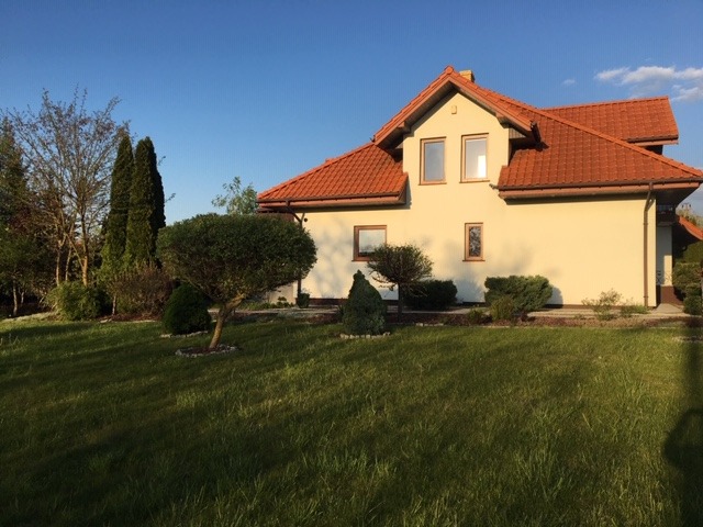 Dwukondygnacyjny budynek z jasną elewacją i czerwoną dachówką, otoczony zielonym trawnikiem z ozdobnymi krzewami i drzewami iglastymi w słoneczny dzień.