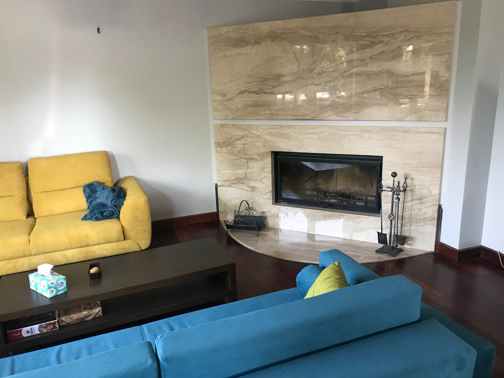 Jasny salon z żółtą sofą, niebieską kanapą i marmurowym kominkiem z czarnym wkładem kominkowym oraz zestawem narzędzi kominkowych.