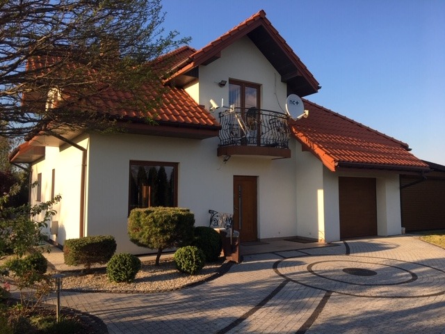 Biały dom jednorodzinny z brązowym dachem i balkonem z kutą balustradą, widoczny z zewnątrz, z zadbanym ogrodem i brukowanym podjazdem z dekoracyjnym wzorem.