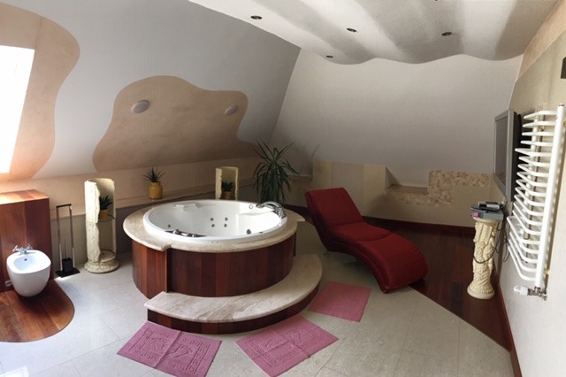 Jasne wnętrze pokoju z jacuzzi otoczonym drewnianą obudową, czerwony leżak, bidet i dekoracyjne kolumny z roślinami.