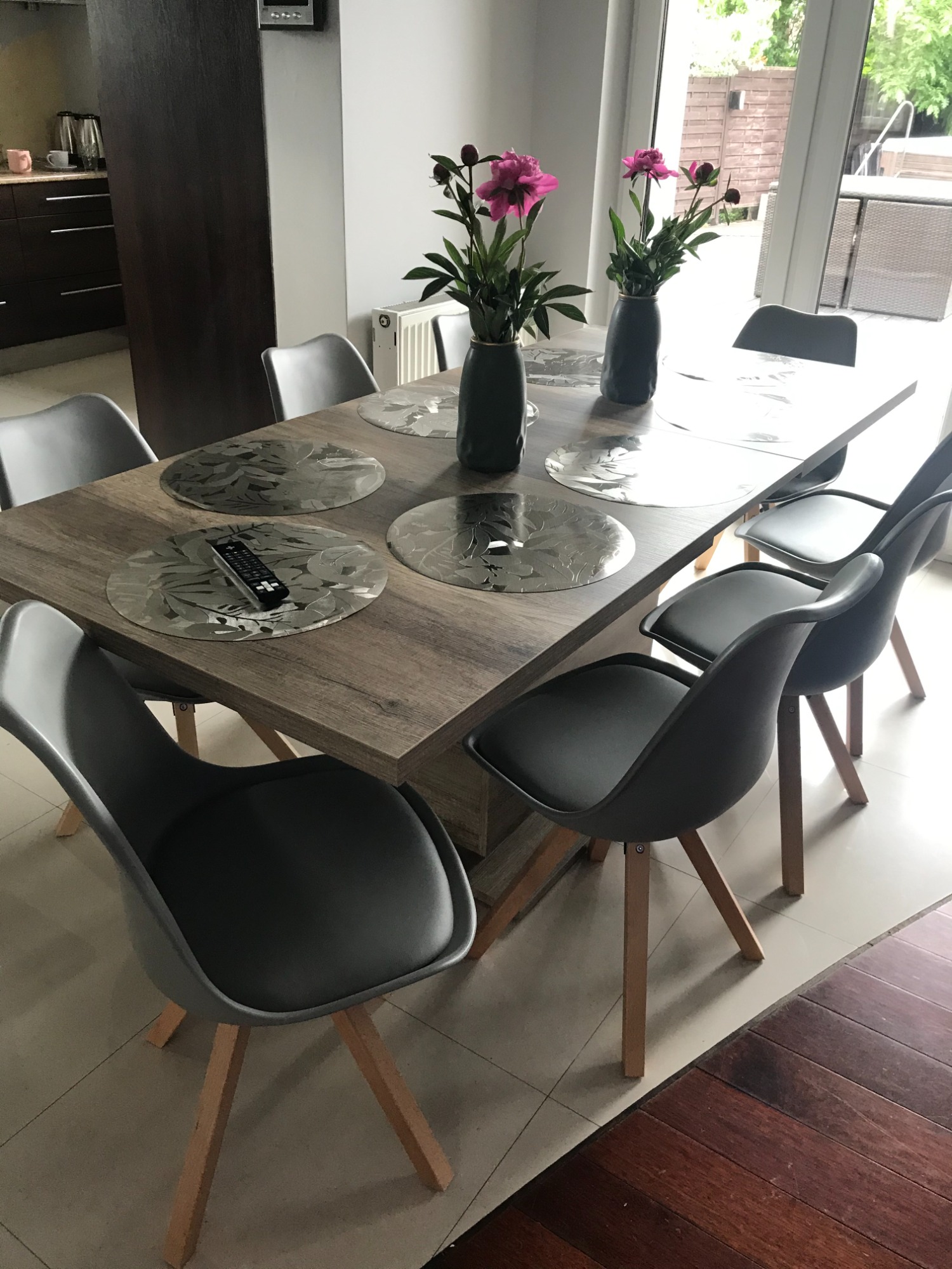 Drewniany stół jadalny z szarymi krzesłami, kwiatami w wazonach i pilotem na podkładce.