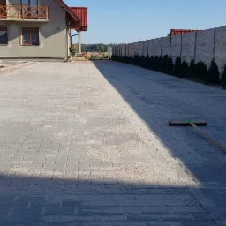 Nowo ułożona, szara kostka brukowa na podjeździe do domu z numerem 53, częściowo zasypana piaskiem, z widoczną miotłą i betonowym ogrodzeniem obsadzonym tujami w tle.