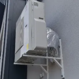 Zewnętrzna jednostka klimatyzacji marki Gree zamontowana na wsporniku ściennym, z folią ochronną na rurach, przy szarej elewacji budynku.