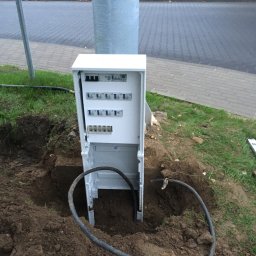 Robim Good Elektroinstalacje - Otwarta, biała skrzynka elektryczna z zainstalowanymi bezpiecznikami i okablowaniem, umieszczona w wykopie ziemnym obok słupa oświetleniowego i chodnika z kostki brukowej.