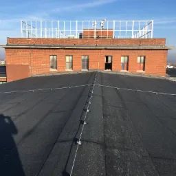 Widok z góry na płaski dach budynku przemysłowego pokryty papą, z widocznym cieniem osoby robiącej zdjęcie i elementami instalacji odgromowej. W tle ceglany budynek z oknami i białą konstrukcją...