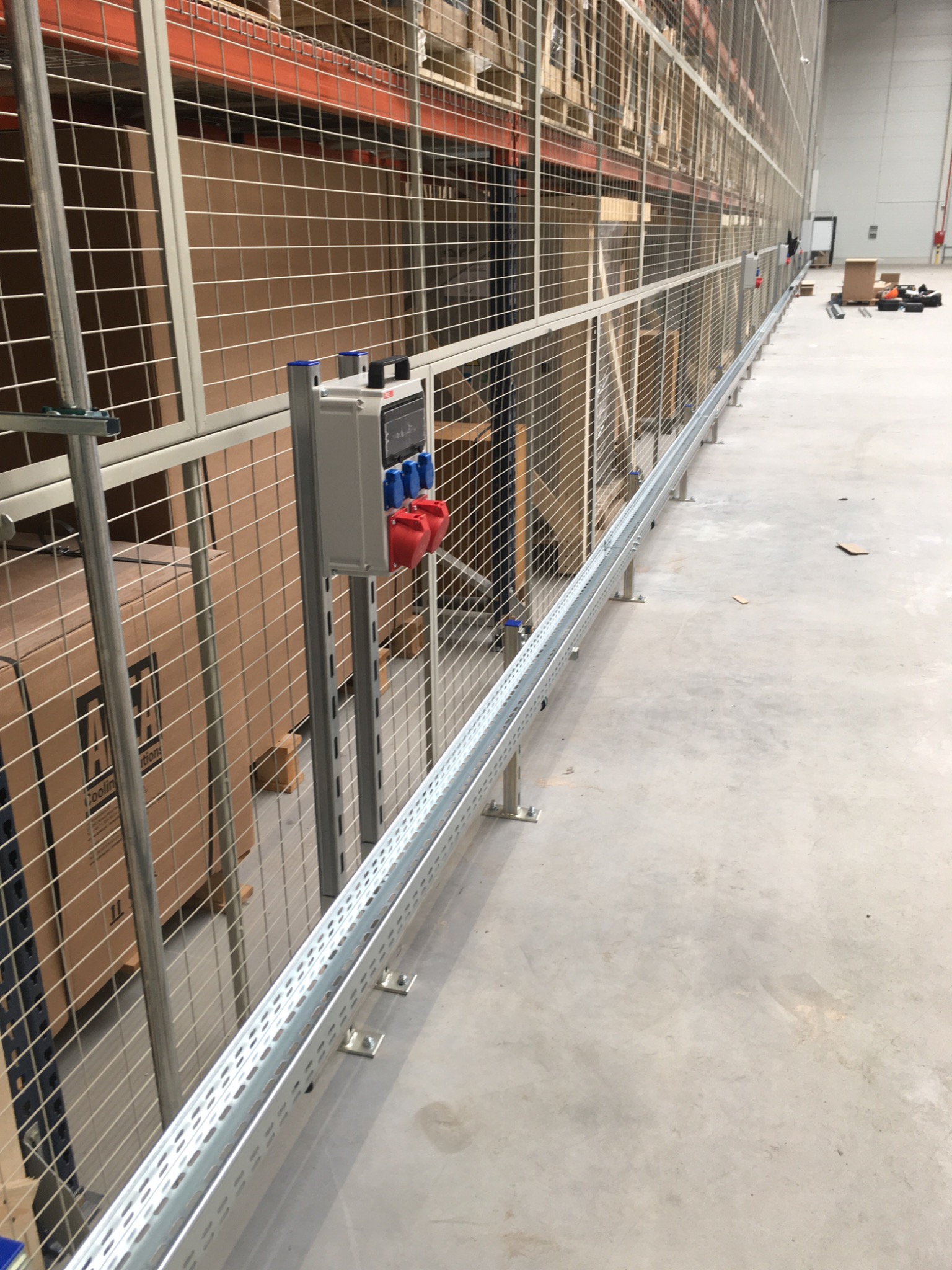 Szyna kablowa z gniazdami siłowymi zamontowana wzdłuż siatki zabezpieczającej regały magazynowe w hali.