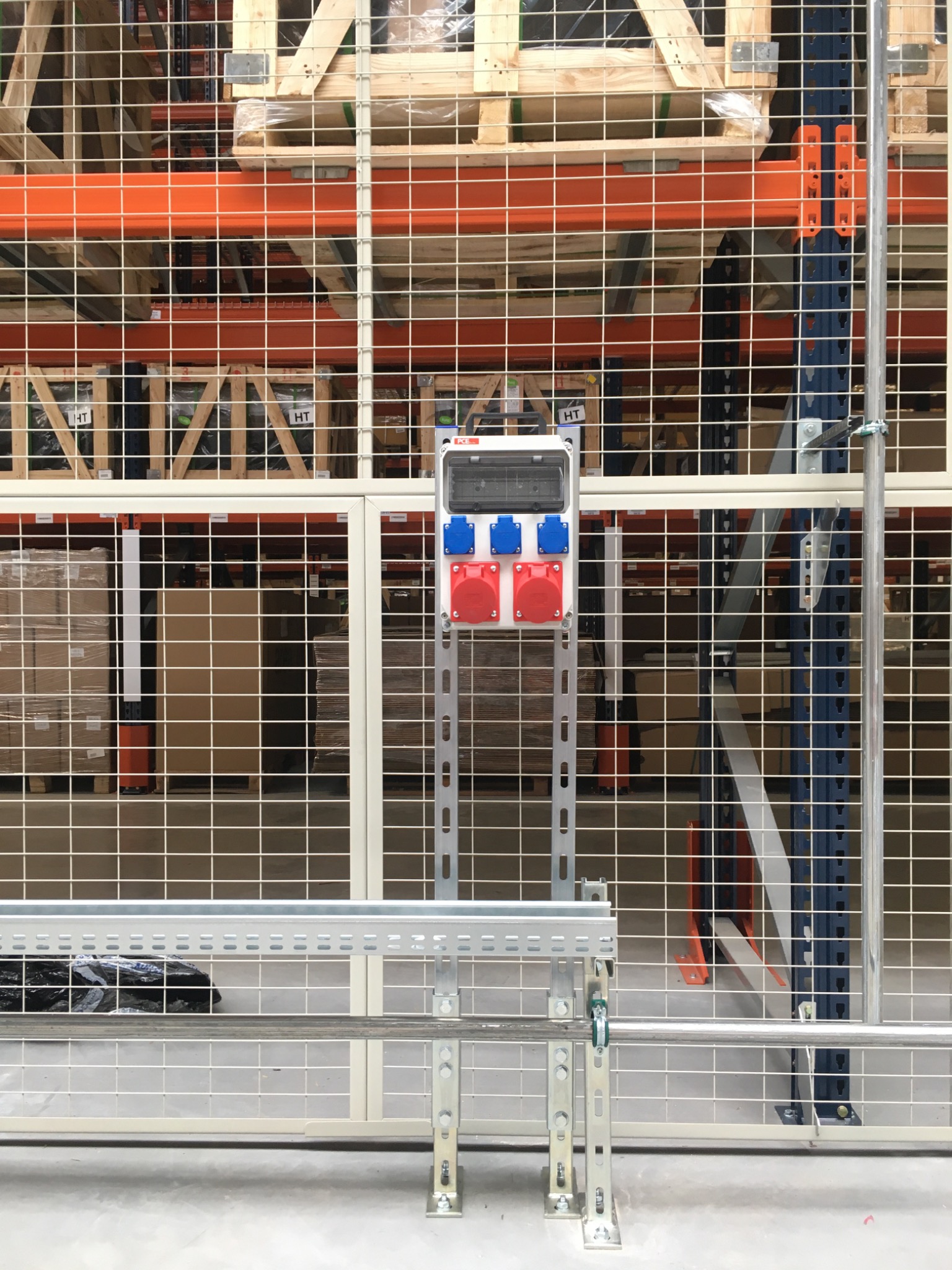 Skrzynka z gniazdami siłowymi zamontowana na metalowej konstrukcji w hali magazynowej we Wrocławiu, widoczna przez siatkę zabezpieczającą.