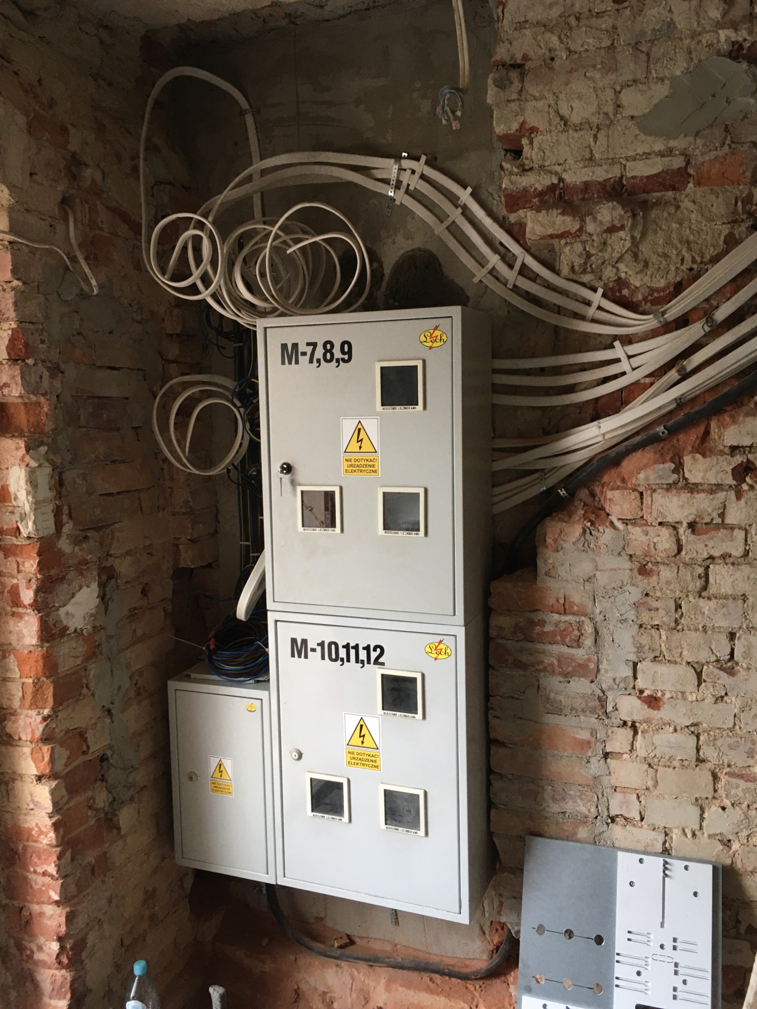 Dwa szare panele elektryczne z zabezpieczeniami i oznaczeniami, zamontowane na surowej ceglanej ścianie w trakcie prac instalacyjnych, z widocznymi przewodami i elementami montażowymi.